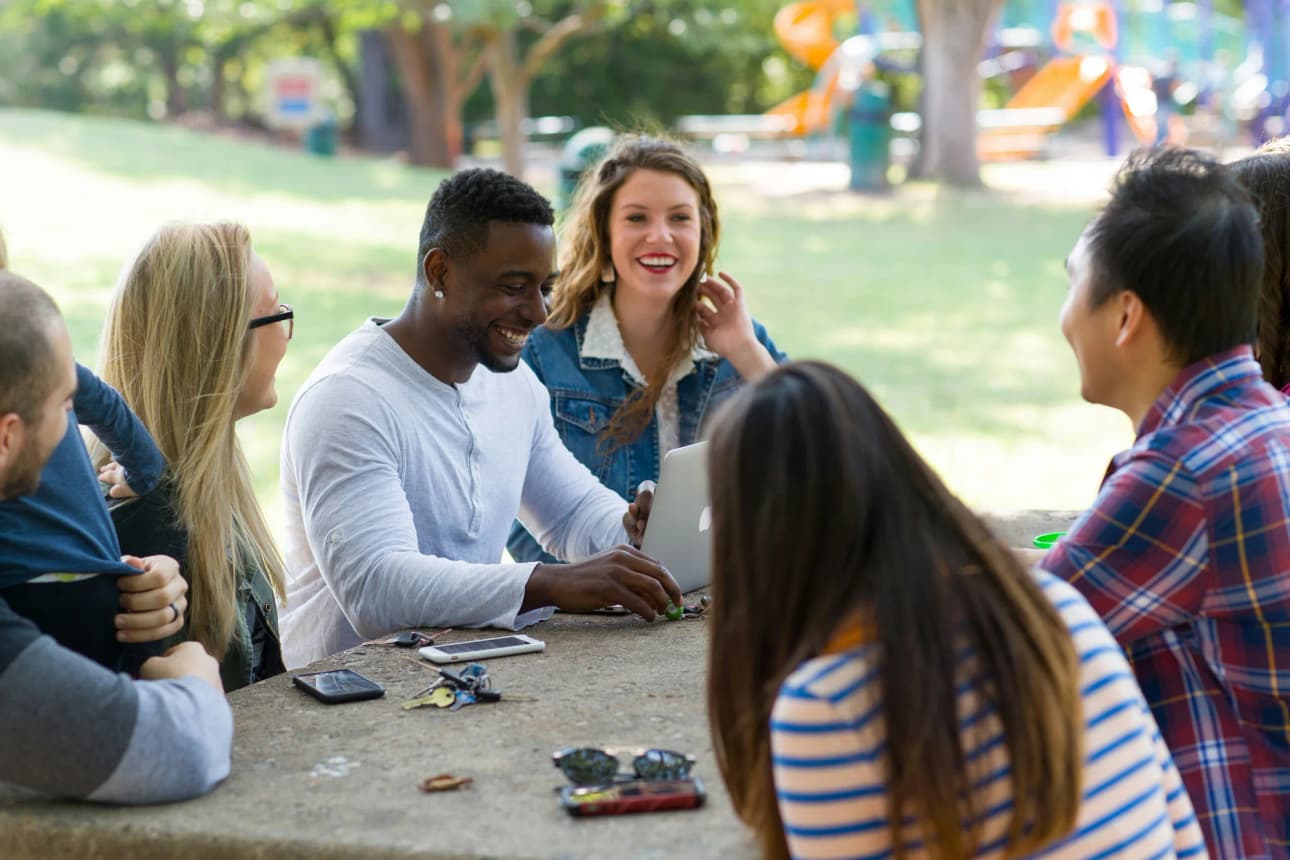 A group of friends gathered around a picnic table in a park, laughing mid-conversation, an open laptop between them