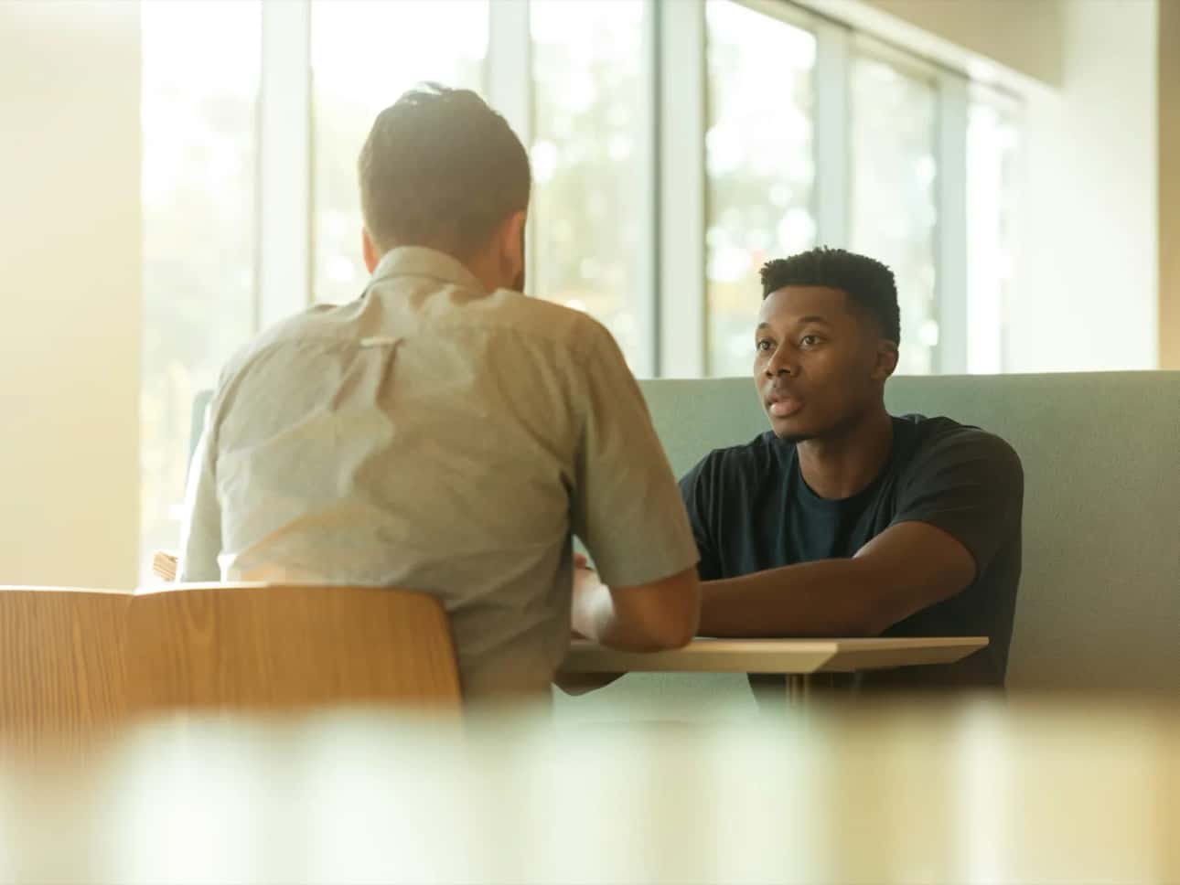 Two men across a booth table in earnest conversation, window light behind them