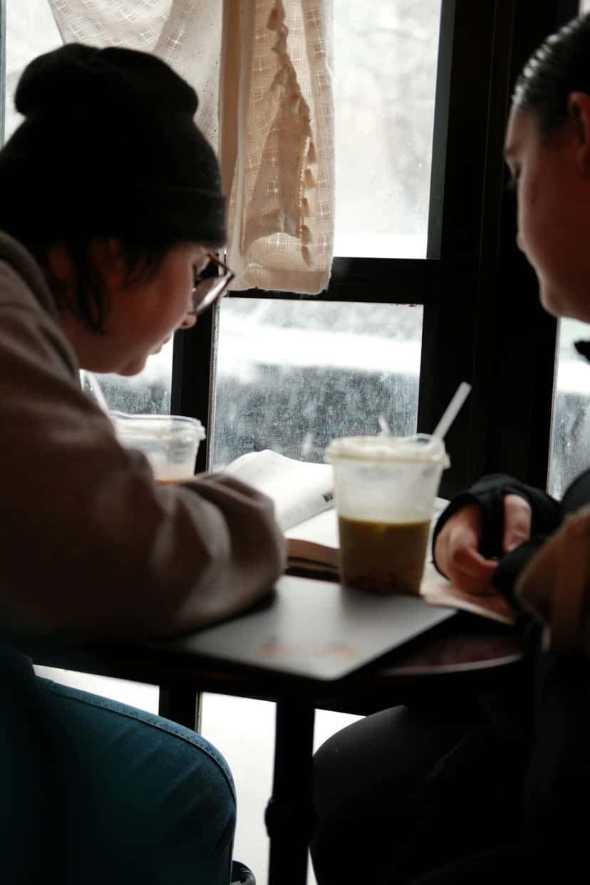 Two friends at a cafe window reading an open book together, warm winter light through frosted glass, iced drinks on the table